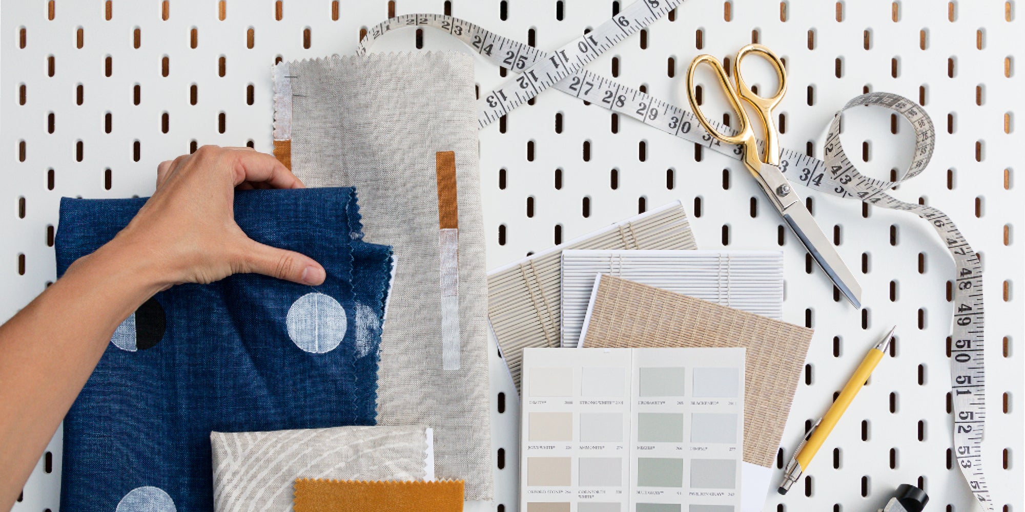 Hand holding collection of fabric samples, beside paint swatches, scissors and measuring tape.