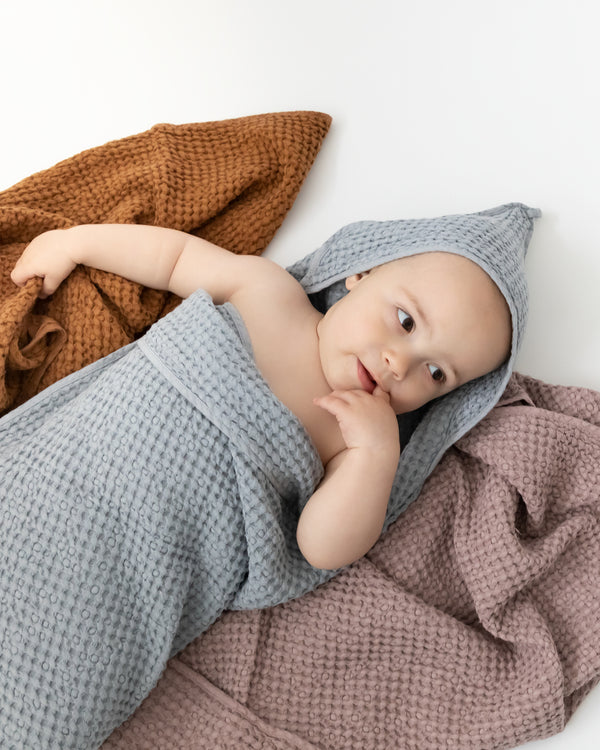 Baby wearing hooded waffle towel in grey blue while laying on top of cinnamon and woodrose coloured towels.