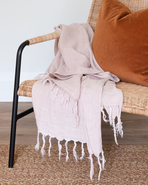 Blush Belgian linen throw with tassels sitting on rattan chair with complimentary rust coloured mohair pillow