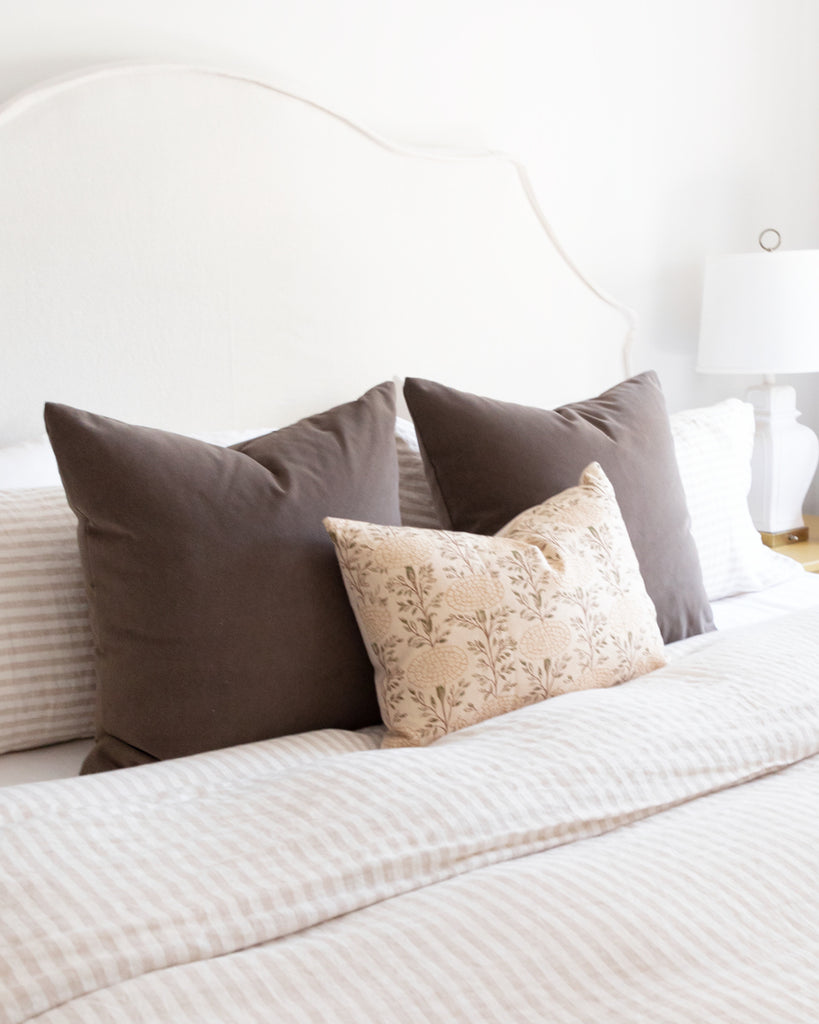 Bedroom setting with two soft brown velvet pillows and beige floral rectangular pillow sitting on a striped duvet 